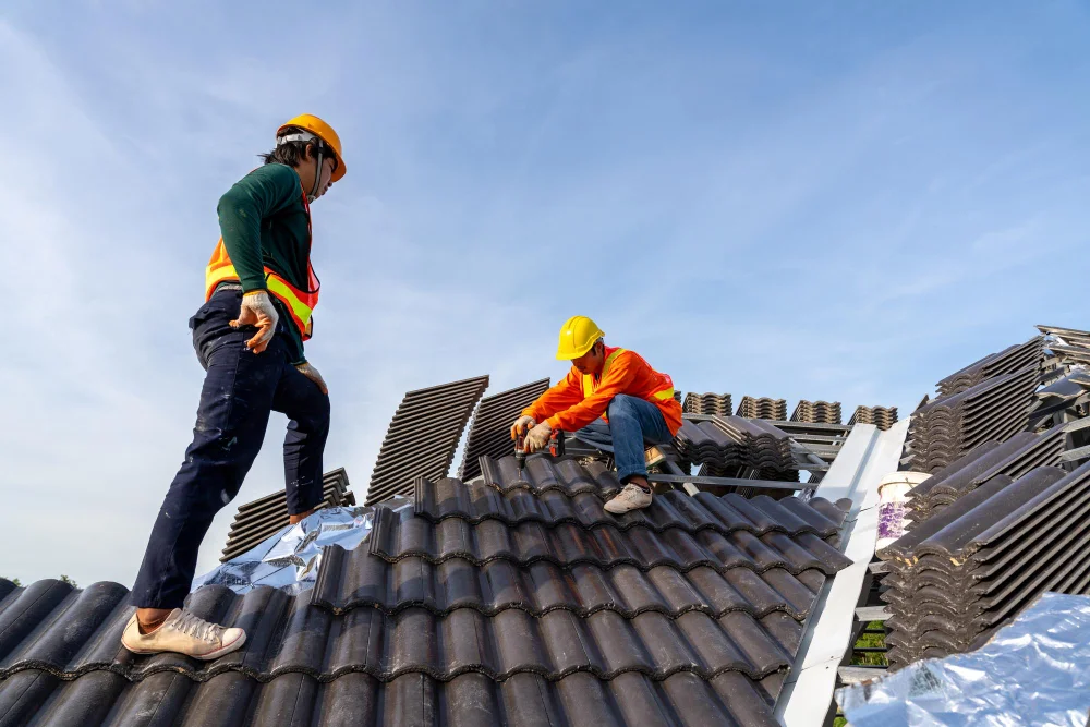 "Two construction workers in safety helmets installing dark ceramic roof tiles on sloped roof"