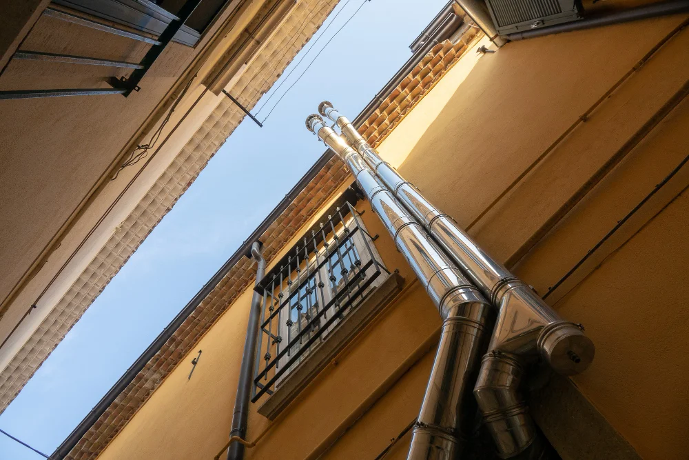 "Tall metal chimney pipes running alongside the exterior wall of a yellow building with a window and blue sky visible above"