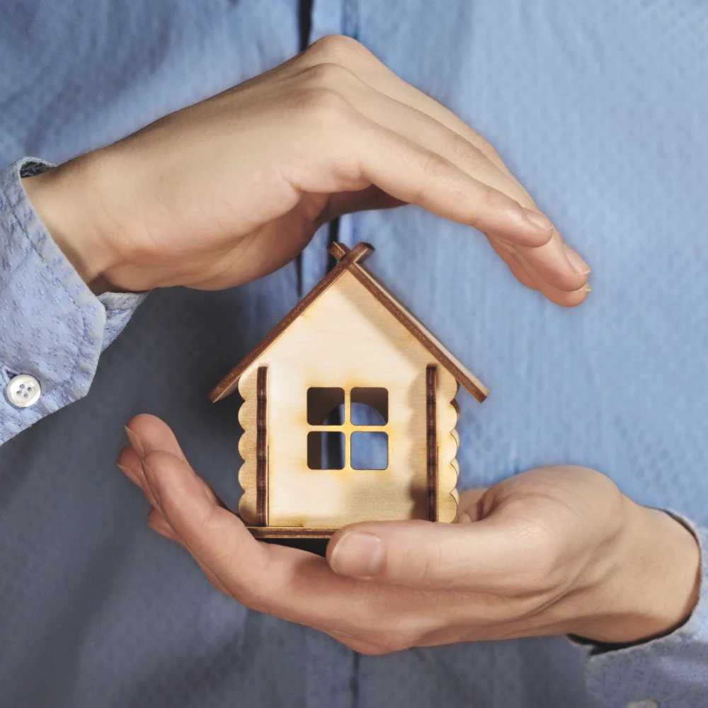 "Hands gently surrounding a wooden house model, symbolizing home safety and care"