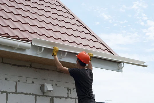 "Construction worker wearing orange safety helmet and gloves installing white rain gutter on the edge of house roof with brown metal tiles"