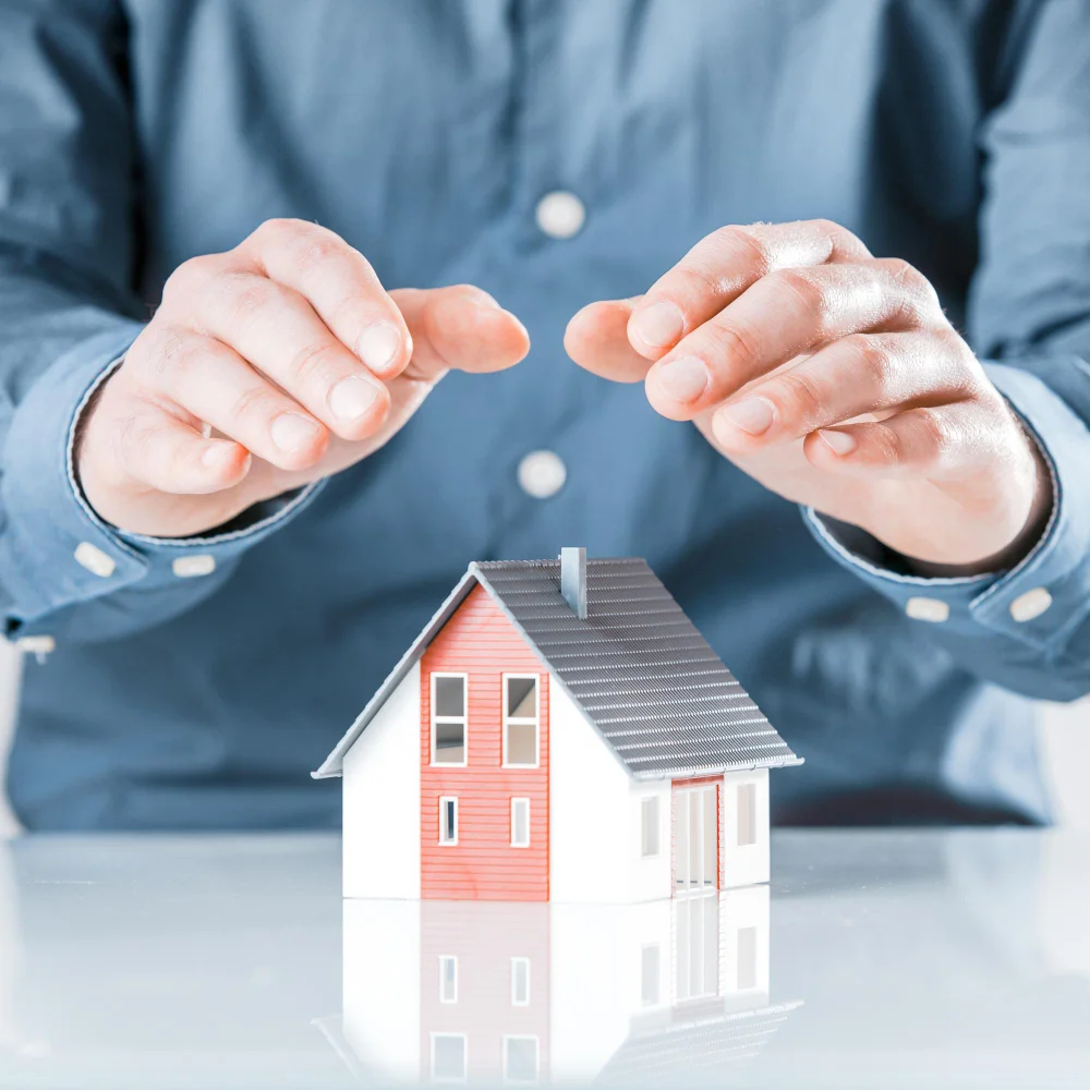 "Person shielding a miniature house model with hands, symbolizing home protection or insurance"