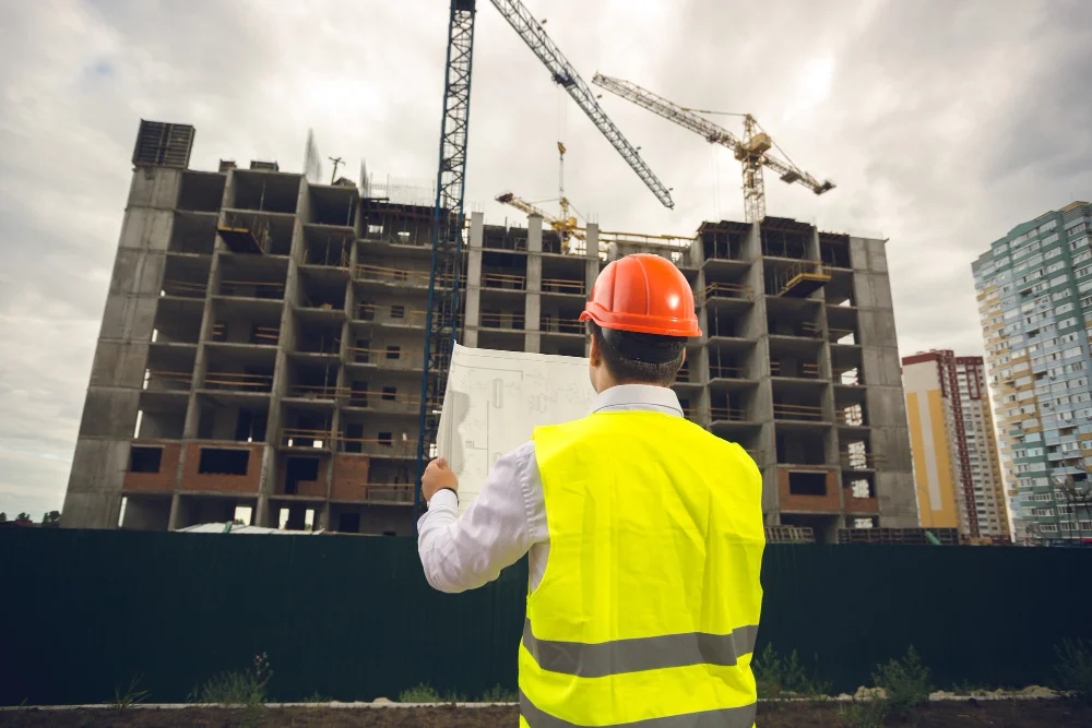 Construction engineer in safety gear reviewing blueprints in front of high-rise building