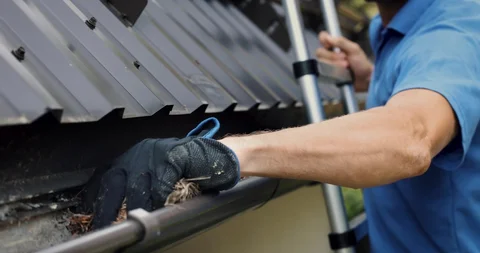 "Worker wearing black glove cleaning debris from a black rain gutter with a blue tool while standing on a ladder"