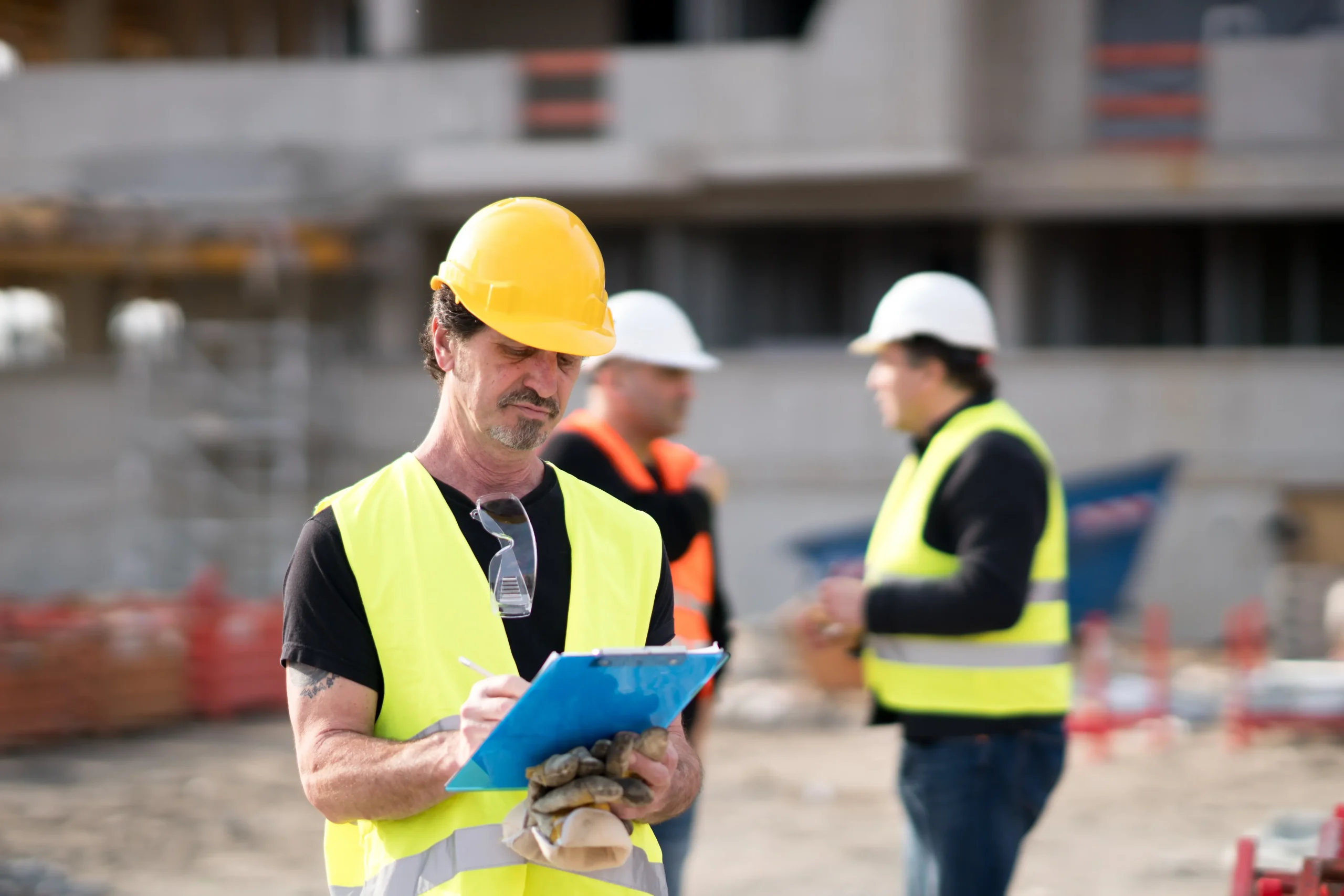 "Construction supervisor in yellow hard hat inspecting site documents with workers in background"