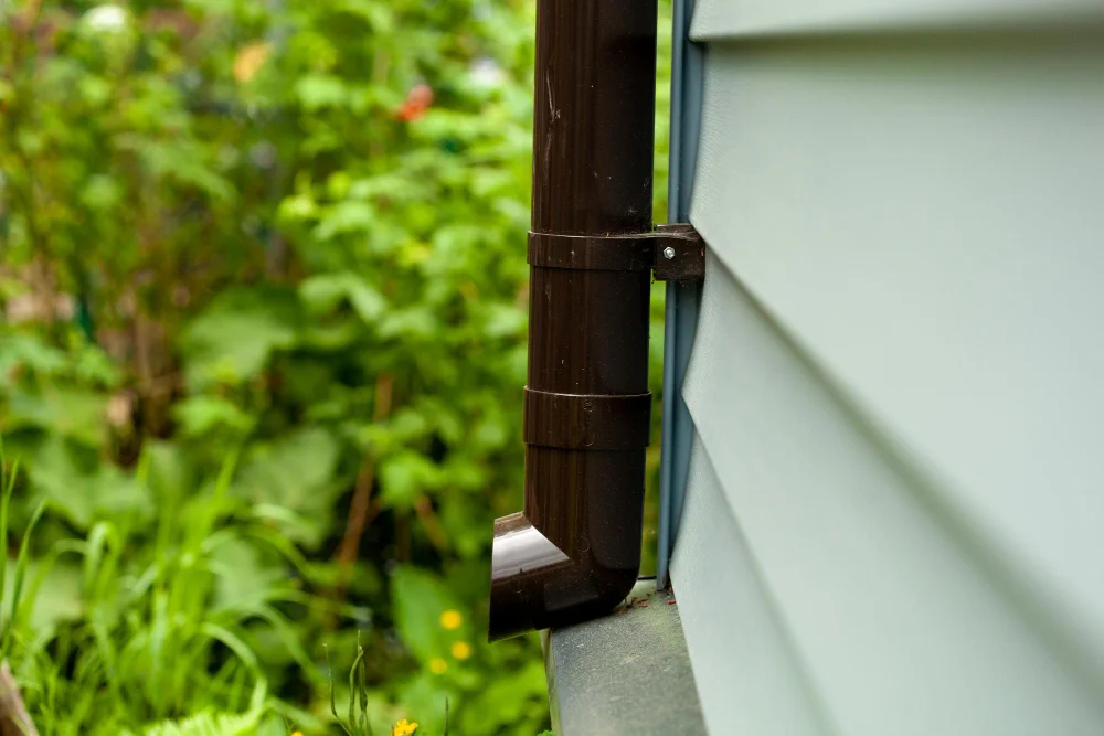 "Close-up of a brown downspout attached to the side of a house with green garden in background"